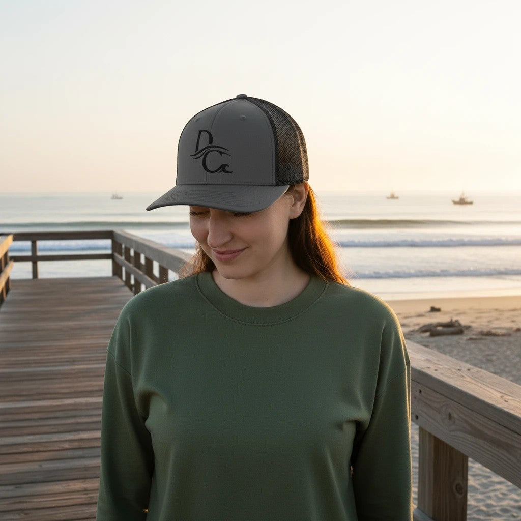 Deepwater Crew Co. grey trucker hat with black mesh back and embroidered DC logo, worn by a woman on a beach boardwalk.