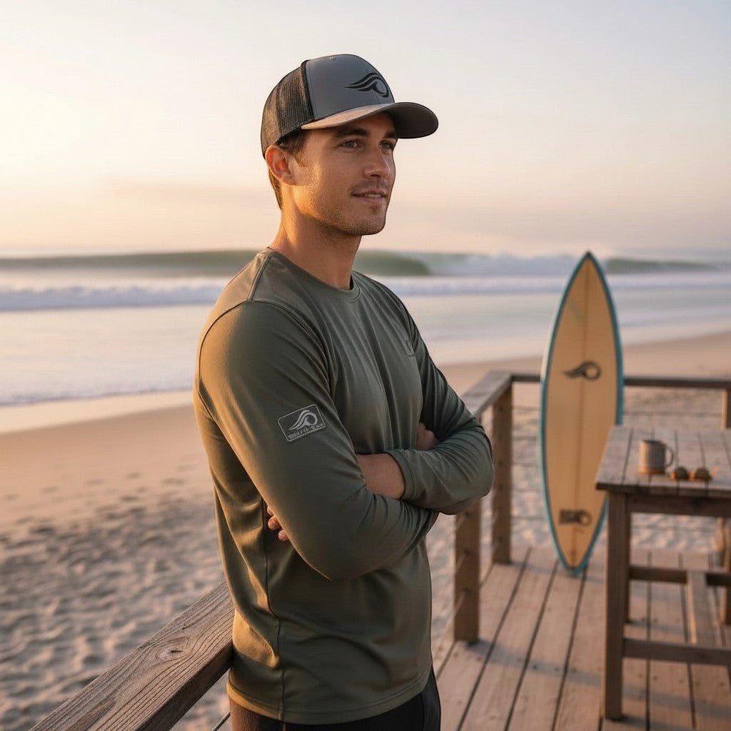 Deepwater Crew Co. grey trucker hat with embroidered wave logo, mesh back, and curved brim. Model on beach.