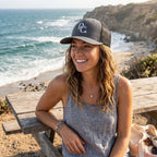 Woman wearing a grey Deepwater Crew Co. trucker hat with a white DC wave logo, outdoors by the ocean.
