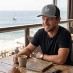 Man wearing a grey and black Deepwater Crew Co. trucker hat with an embroidered wave logo at a seaside cafe.