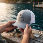 Deepwater Crew Co. grey and white mesh trucker hat with embroidered logo held over a wooden dock by the water.