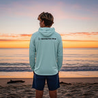 Man wearing a light blue Deepwater Crew Co. performance UPF hoodie standing on a beach at sunset.