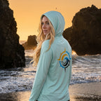 Deepwater Crew Co. light blue performance UPF hoodie with a compass logo on the back, worn by a woman at the beach.