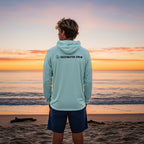 Man wearing a light blue Deepwater Crew Co. UV performance hoodie standing on a beach at sunset.