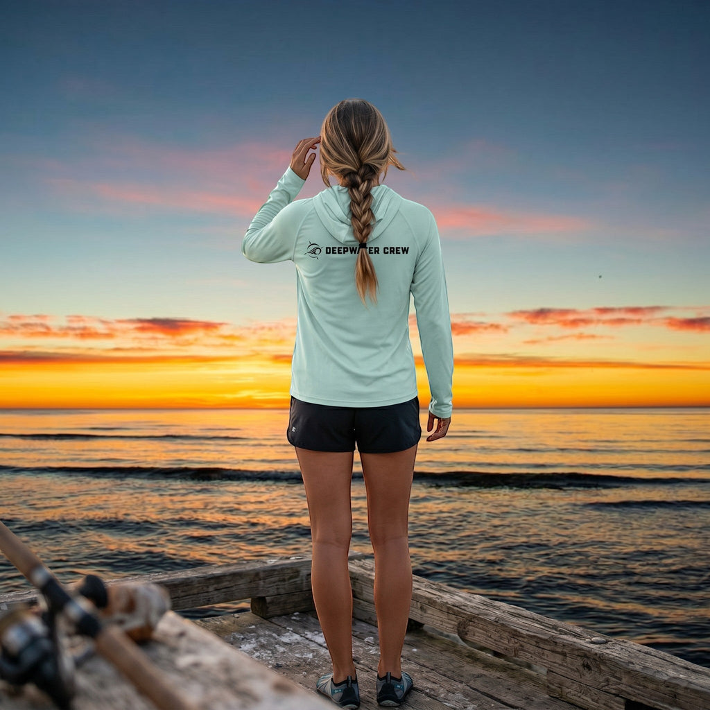 Woman wearing a light green Deepwater Crew Co. UPF performance hoodie standing on a dock at sunset.