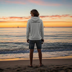 Man wearing a light grey Deepwater Crew Co. performance UPF hoodie standing on a beach at sunset.
