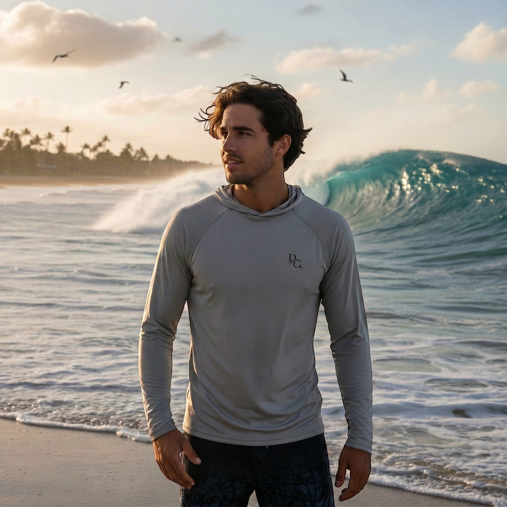 A man wearing a light grey Deepwater Crew Co. UPF performance hoodie on a beach with a large ocean wave in the background