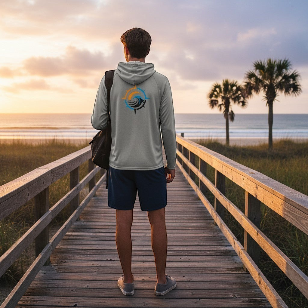 Man wearing a light grey Deepwater Crew Co. UPF performance hoodie with a compass logo on a wooden boardwalk at sunset.