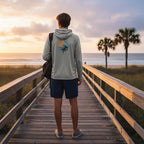 Man wearing a light grey Deepwater Crew Co. UPF performance hoodie with a compass logo on a wooden boardwalk at sunset.
