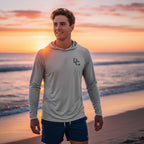Man wearing a Deepwater Crew Co. light grey UV performance hoodie on a beach at sunset with raglan sleeves.