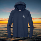 Deepwater Crew Co. navy blue performance UV hoodie with raglan sleeves and white logo against a sunset ocean backdrop