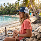 Deepwater Crew Co. navy and white mesh trucker cap with flat bill and embroidered logo worn by a woman on a beach.