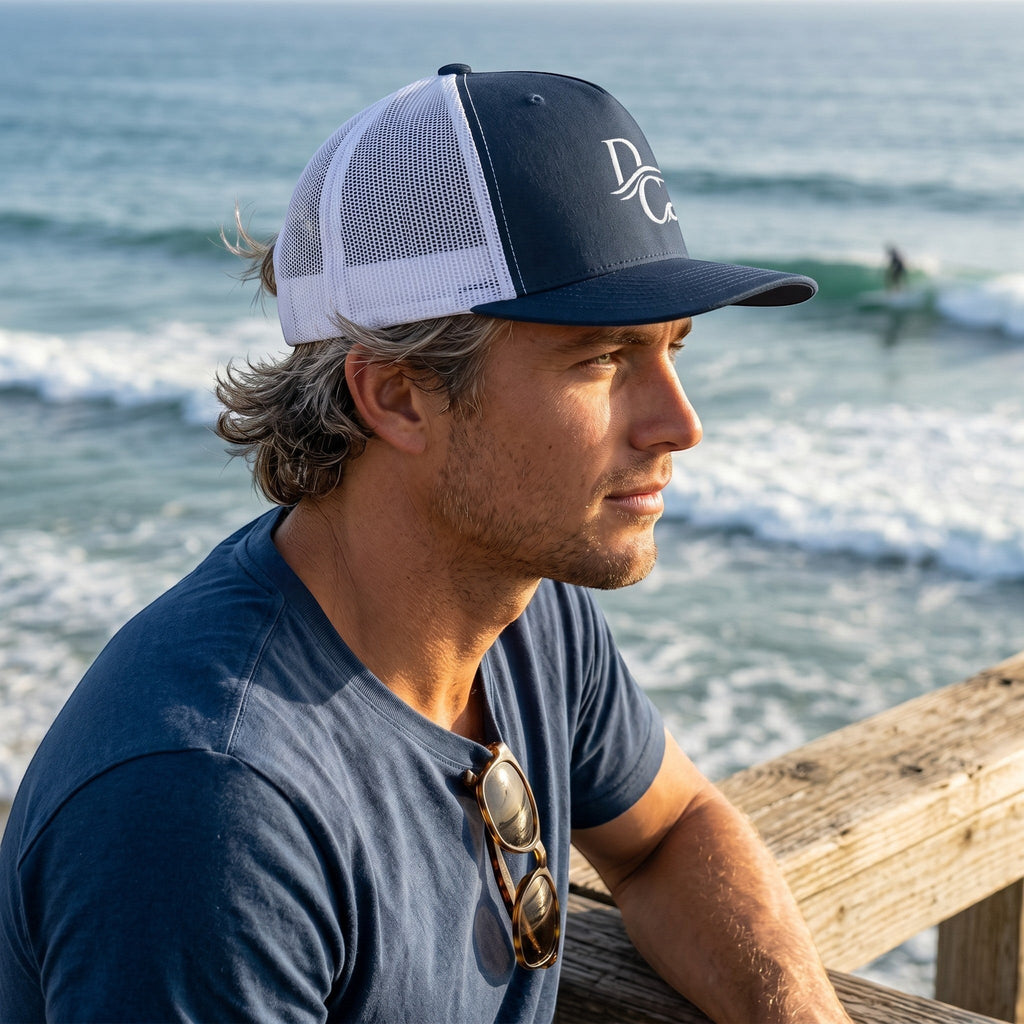 Deepwater Crew Co. navy and white 5-panel trucker cap with embroidered logo worn by a man overlooking the ocean