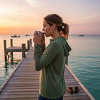 Deepwater Crew Co. woman wearing an olive green tri-blend long sleeve hoodie while drinking coffee on a wooden dock.