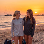 Two women wearing Deepwater Crew Co. UPF 44+ performance hoodies on a beach at sunset with a sailboat in the background.