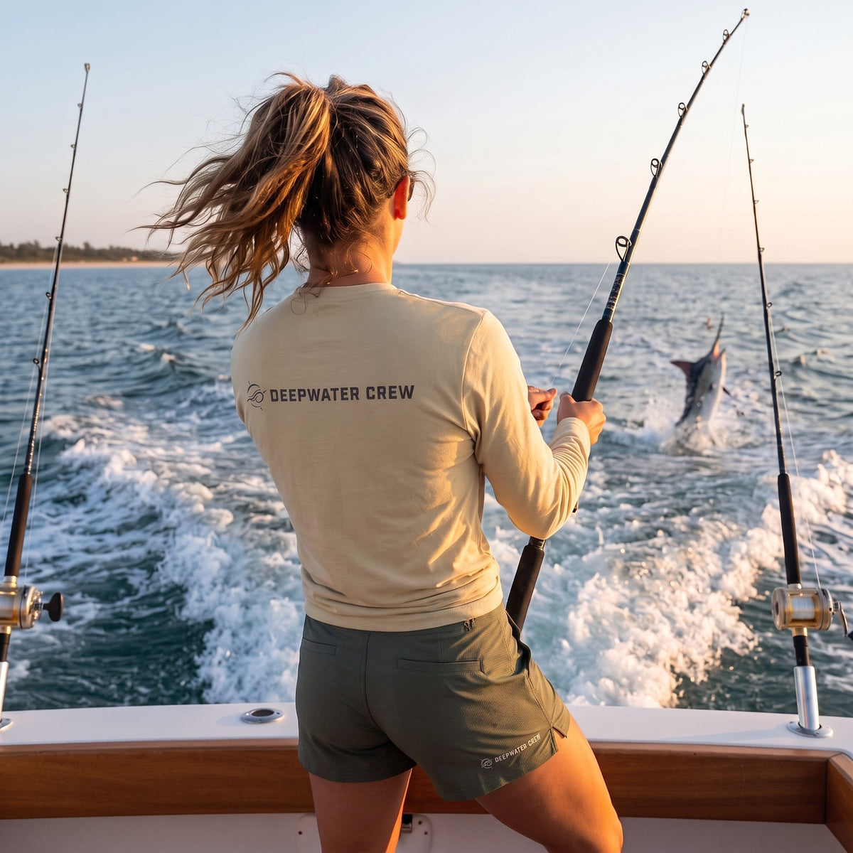Woman wearing a Deepwater Crew Co. tan long-sleeve performance shirt fishing on a boat with a marlin jumping in the sea.