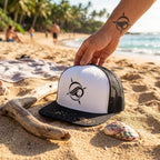 Deepwater Crew Co. white and black trucker cap with flat bill and embroidered logo held on a sunny beach.