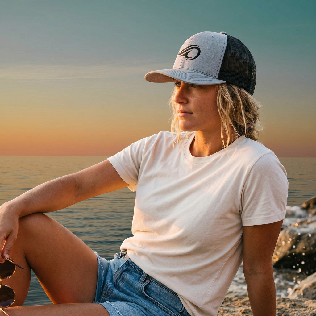 Woman wearing a Deepwater Crew Co. grey and black mesh trucker hat with a wave logo at sunset by the ocean.