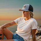 Woman wearing a Deepwater Crew Co. grey and black mesh trucker hat with a wave logo at sunset by the ocean.