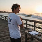 Deepwater Crew Co. white cotton t-shirt with ocean-inspired back graphic worn by a man on a pier at sunset.