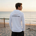 Man wearing a white Deepwater Crew Co. long sleeve hoodie with a logo on the back, standing on a beach at sunset.