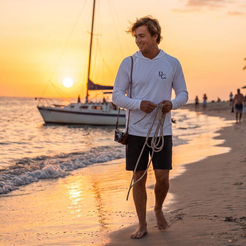 A man wearing a white Deepwater Crew Co. UPF 44+ performance hoodie walking on a beach at sunset with a sailboat.