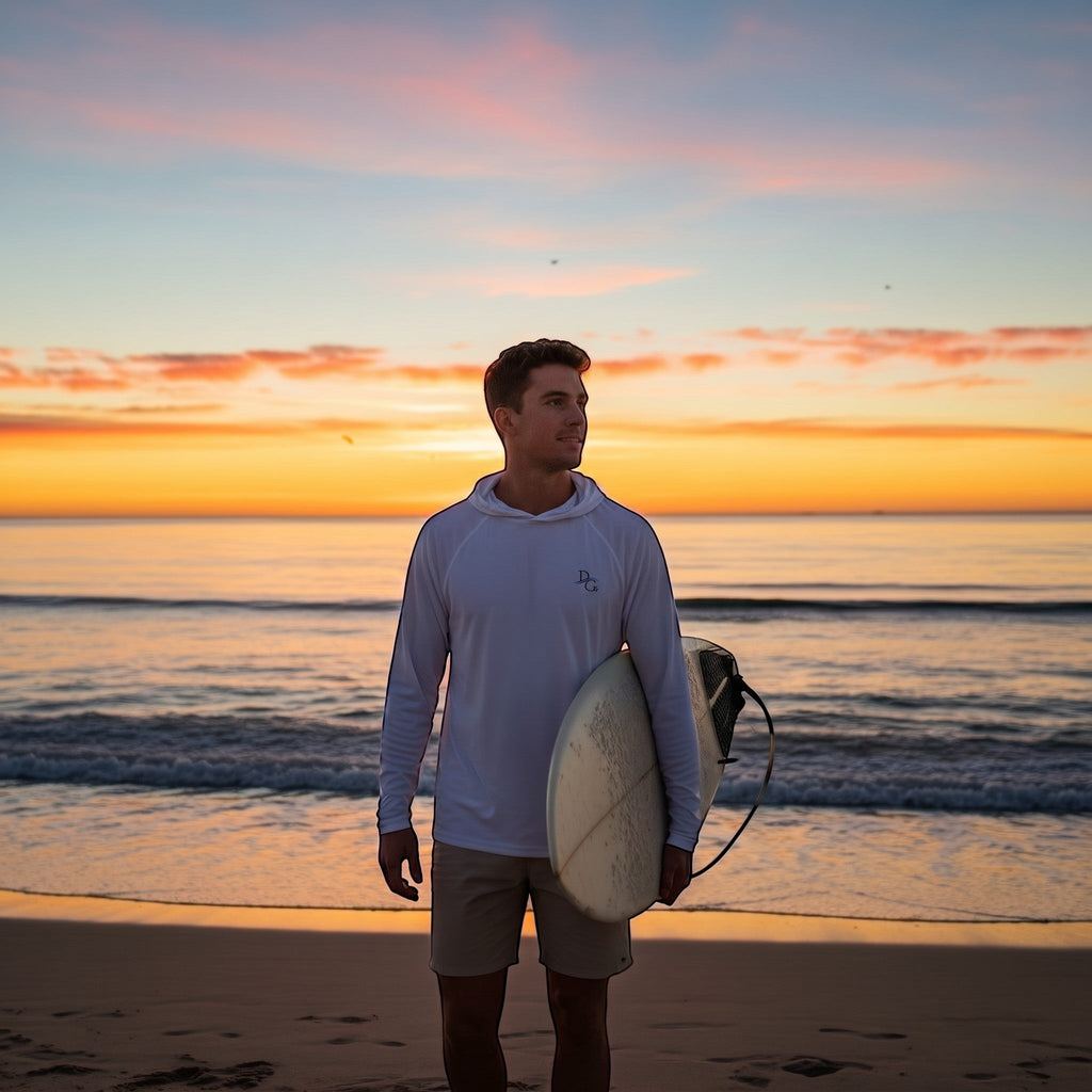Deepwater Crew Co. man wearing a white performance UPF hoodie with a surfboard on the beach at sunset.